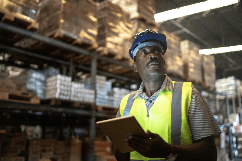 A light industrial staffing hire holds a tablet while surveying shelves in a warehouse.