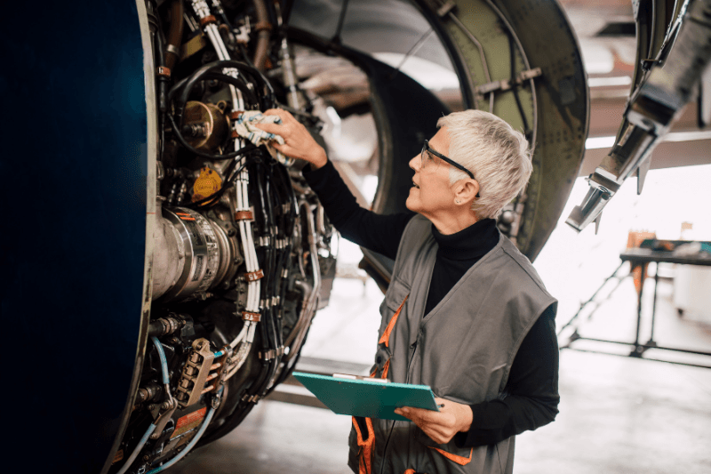 An aerospace recruitment hire inspects mechanical parts of a plane while holding a clipboard.