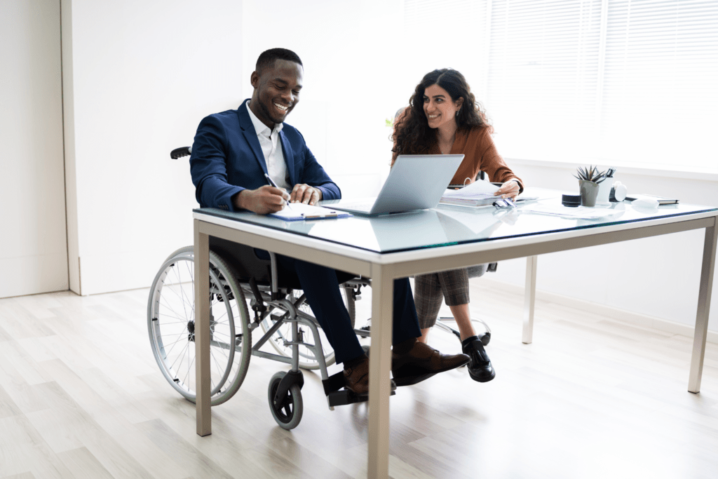 A professional services staffing expert in a wheelchair works at a desk beside another employee while smiling.